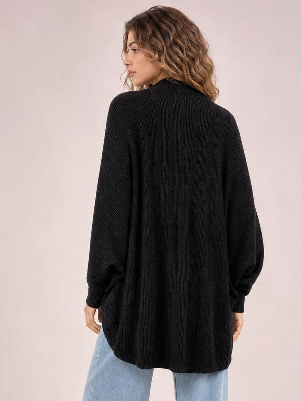 A model stands with her back to the camera wearing a black knit open-front cardigan with a relaxed oversized silhouette, long sleeves, and ribbed cuffs.
