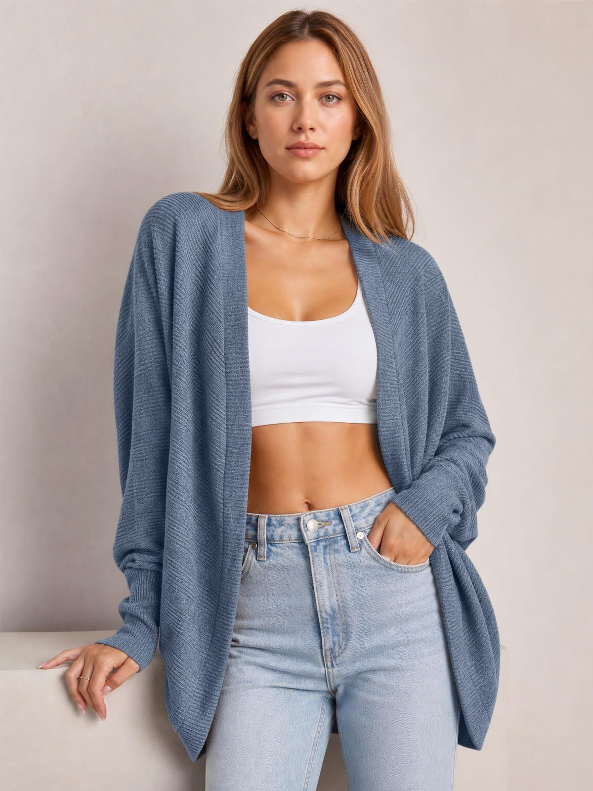 Model wearing a blue knit open front cardigan with long sleeves and a relaxed draped silhouette over a white cropped top and jeans.