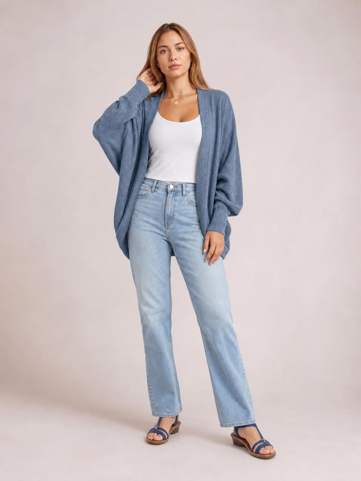 Model poses in a relaxed stance wearing a blue knit open-front cardigan with a long, loose silhouette, ribbed trim, and dropped sleeves over a white tank top.