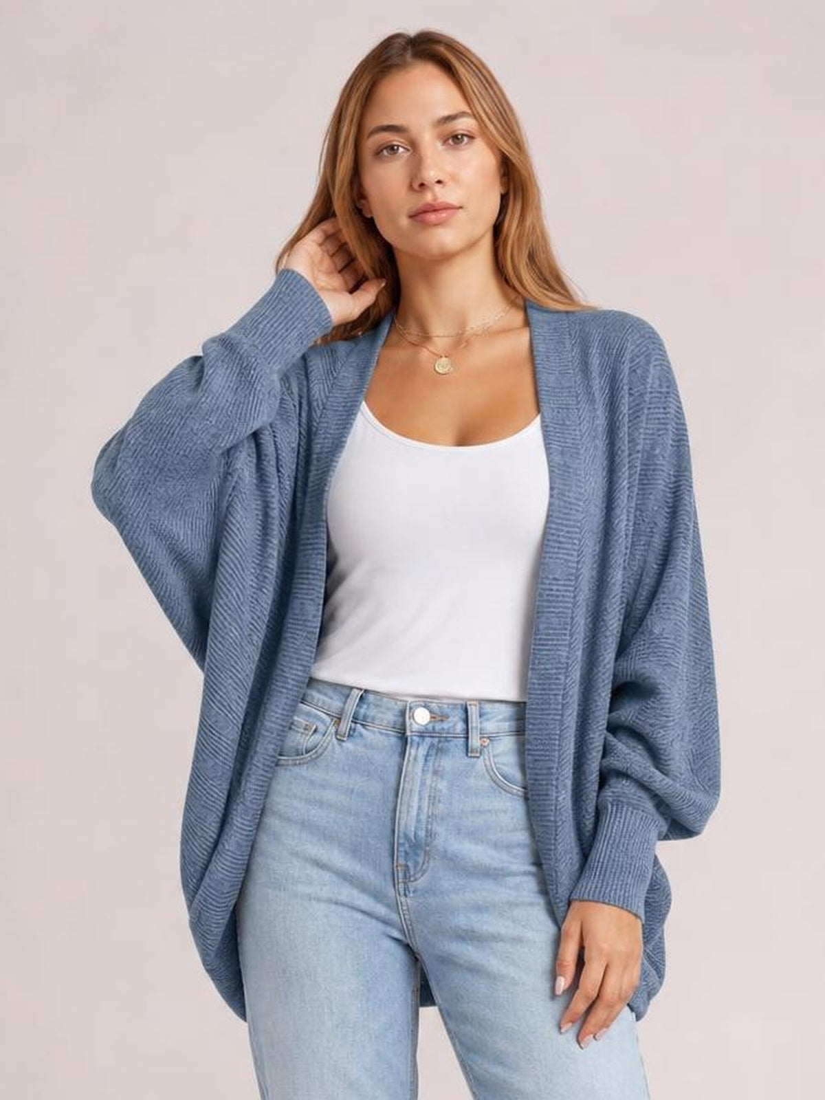 Model poses in a blue knit open front cardigan with a relaxed oversized silhouette, long sleeves, ribbed cuffs, and a white tank top.