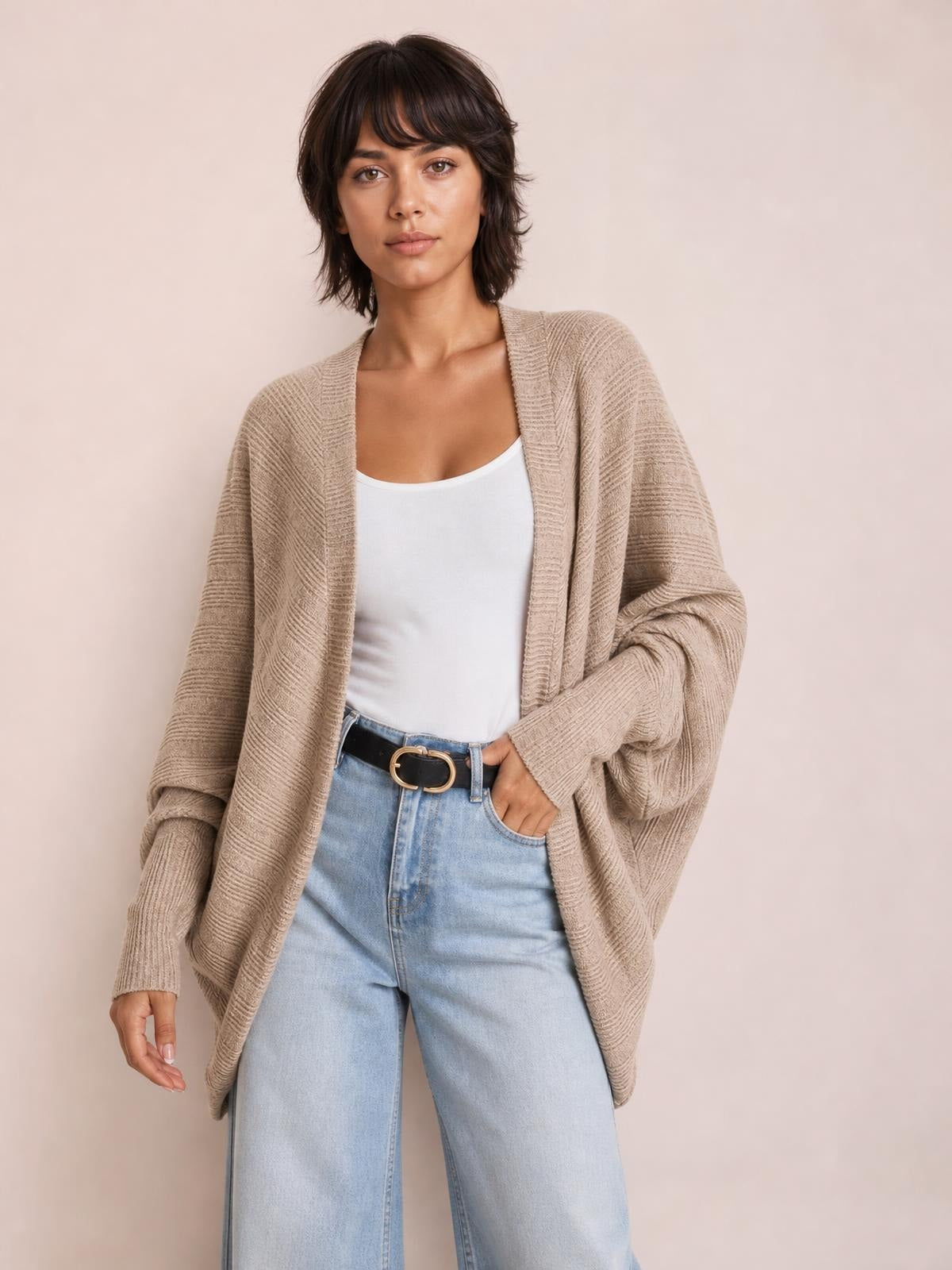 A model poses with one hand in her jeans pocket, wearing a taupe textured knit open-front cardigan with a relaxed longline silhouette and sleeves.