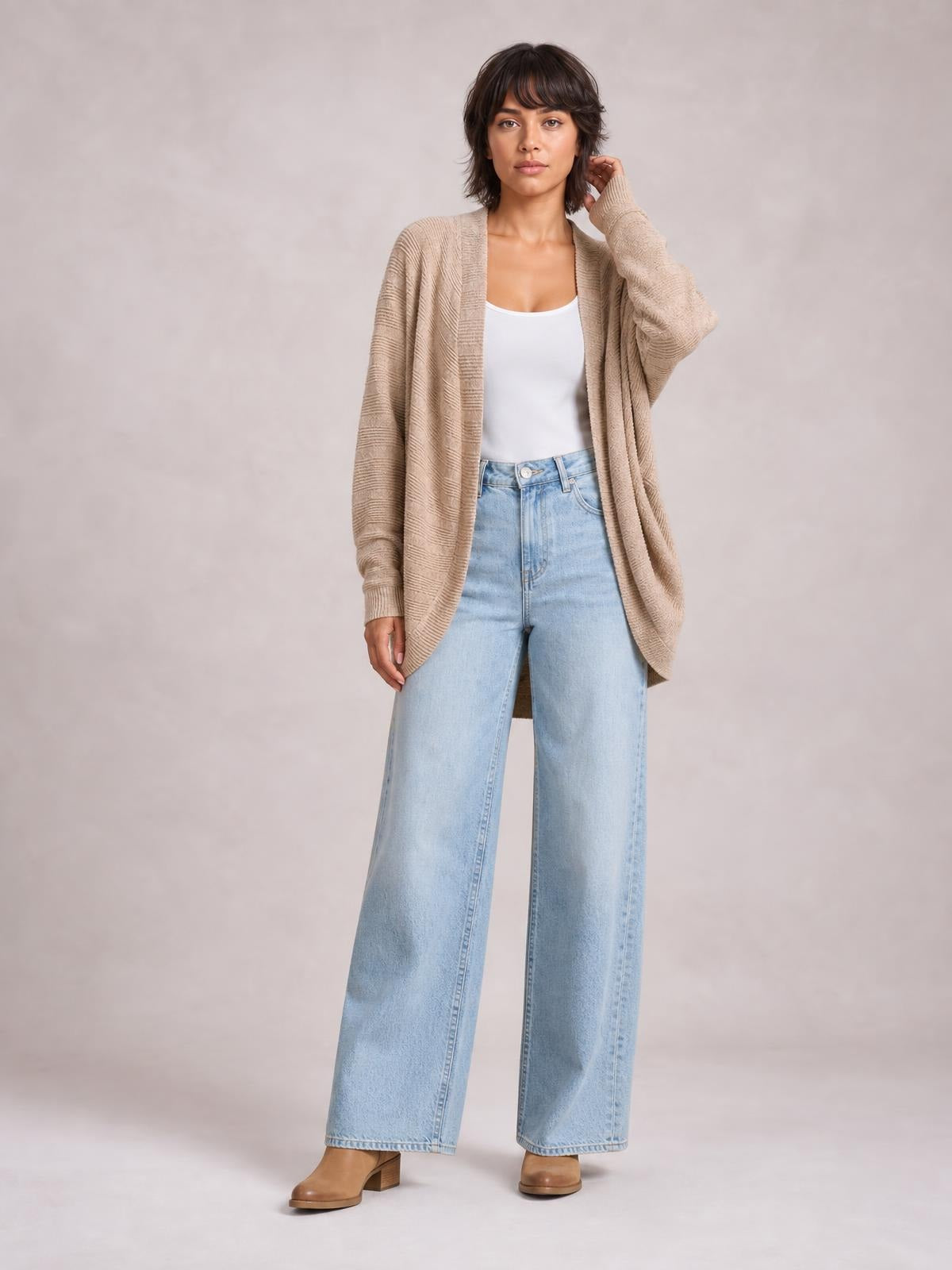 Model wears a beige knit open-front cardigan with a relaxed longline silhouette, textured sleeves, and an easy drape over a white tank and jeans.