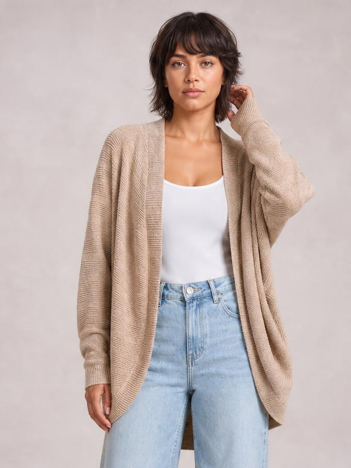 A model wears a beige knit open-front cardigan with a relaxed longline silhouette, textured construction, and ribbed cuffs over a white top and jeans.
