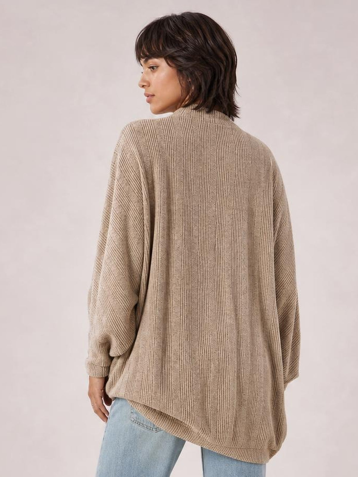 Model shown from behind in a relaxed pose wearing a taupe knit open-front cardigan with an oversized silhouette, ribbed vertical texture, long sleeves, and ribbed cuffs and hem.
