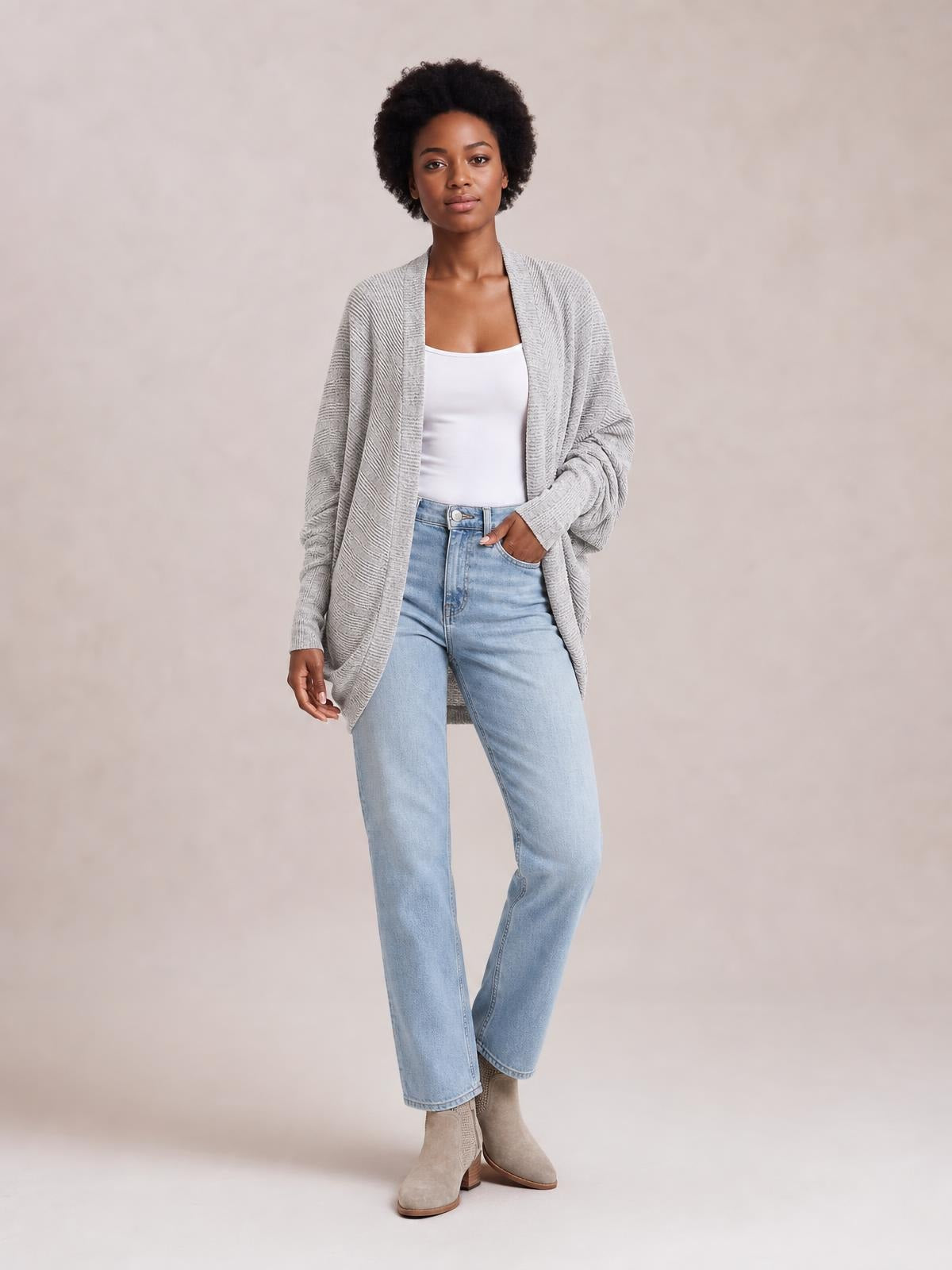 Model stands in light-wash jeans wearing a gray knit open front cardigan with a relaxed, longline silhouette and subtle textured construction.