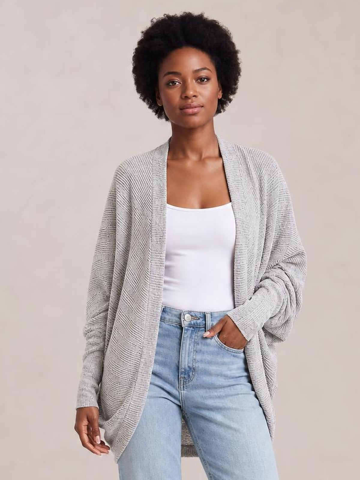 Model wearing a light gray knit open front cardigan with a relaxed longline silhouette, draped sleeves, and ribbed trim over a white top and jeans.