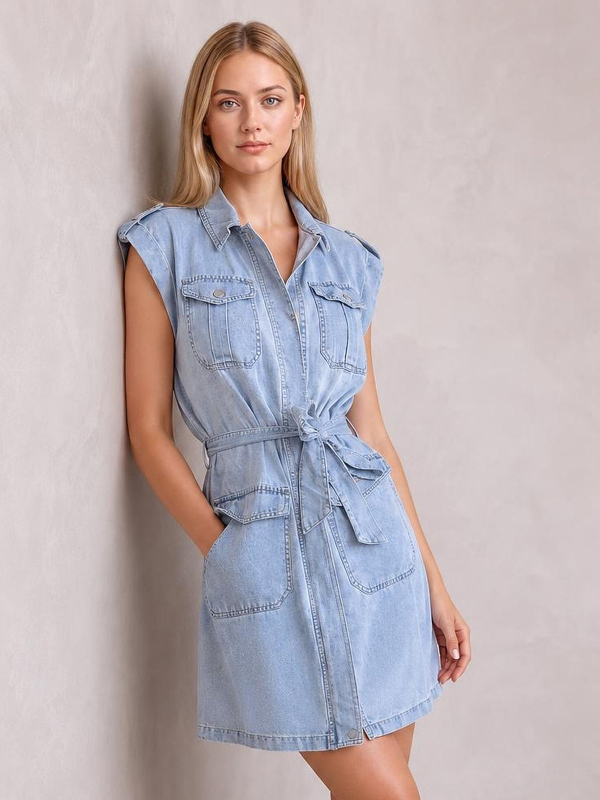 A model poses in a light blue denim mini dress with a collared button front, rolled sleeves, utility pockets, and a tied waist belt.