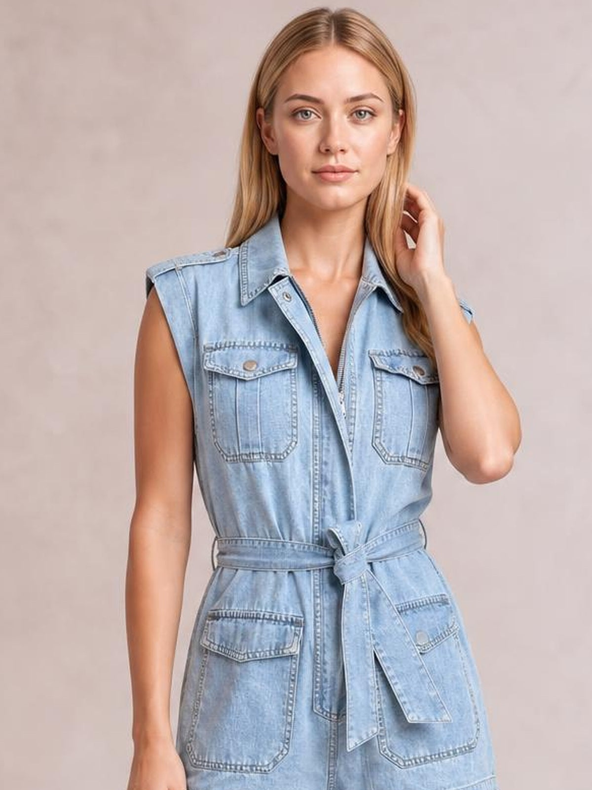 A model poses in a light blue denim mini dress with a sleeveless collared design, button front, multiple flap pockets, and a tied waist belt.