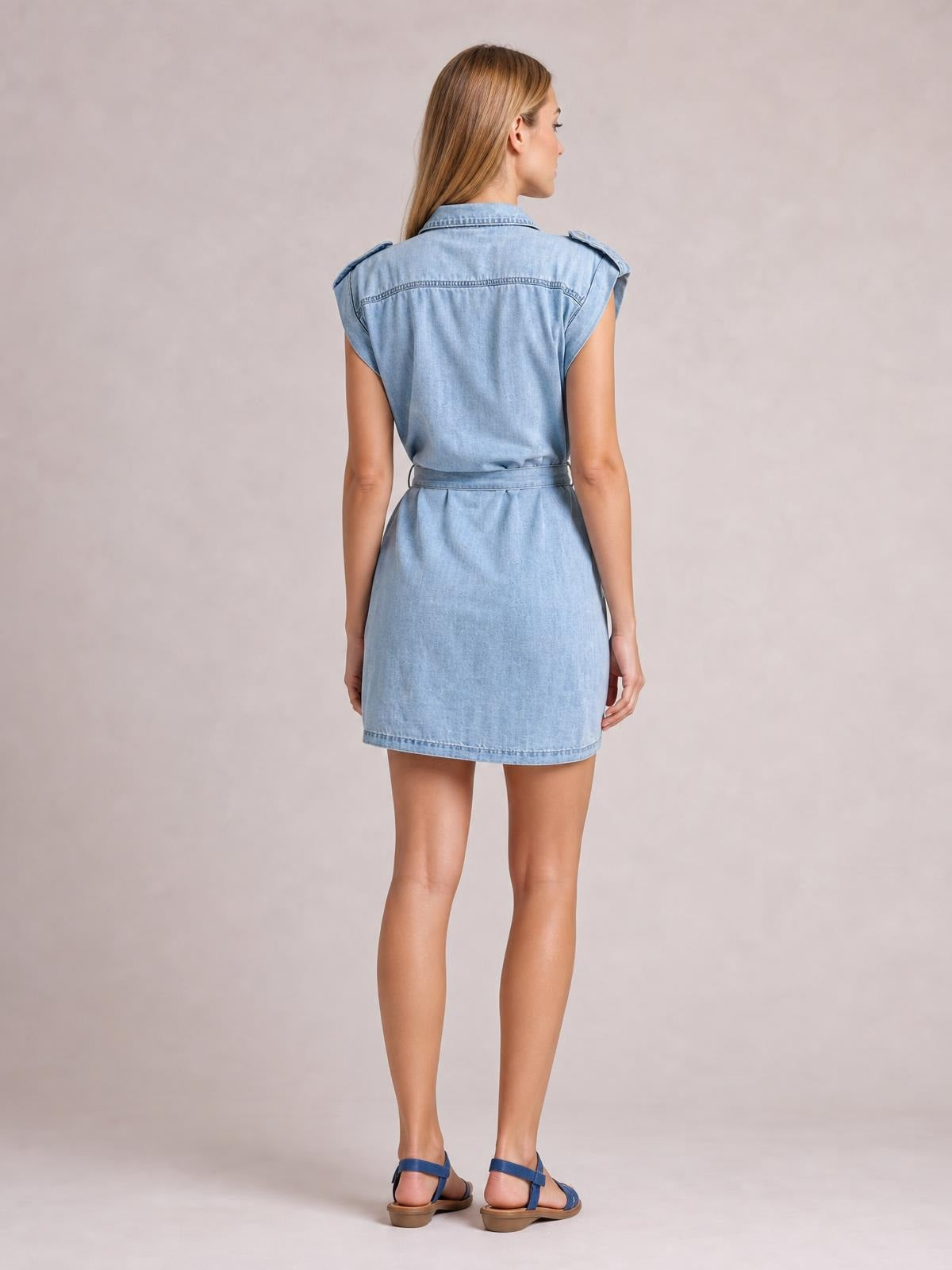 Model shown from behind in a light blue denim mini dress with a collared neckline, sleeveless rolled shoulders, waist tie belt, and straight silhouette.