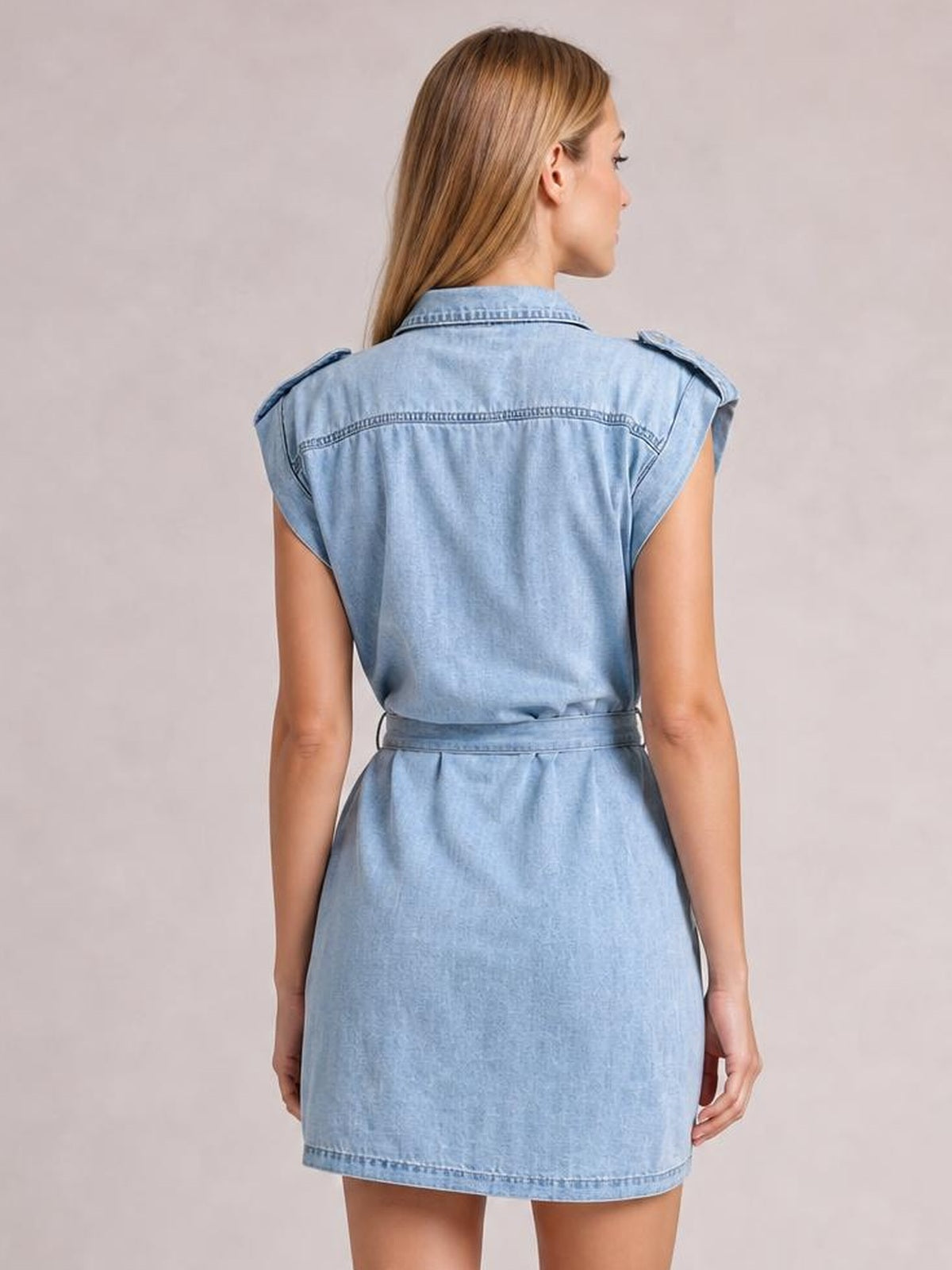 Model shown from behind in a light blue denim mini dress with a collared neckline, sleeveless cut, shoulder tabs, stitched back yoke, and tie waist belt.