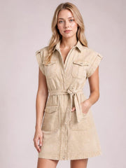 A model stands facing forward in a light beige denim mini dress with a collared neckline, sleeveless cut, tied waist, utility pockets, and an A-line silhouette.