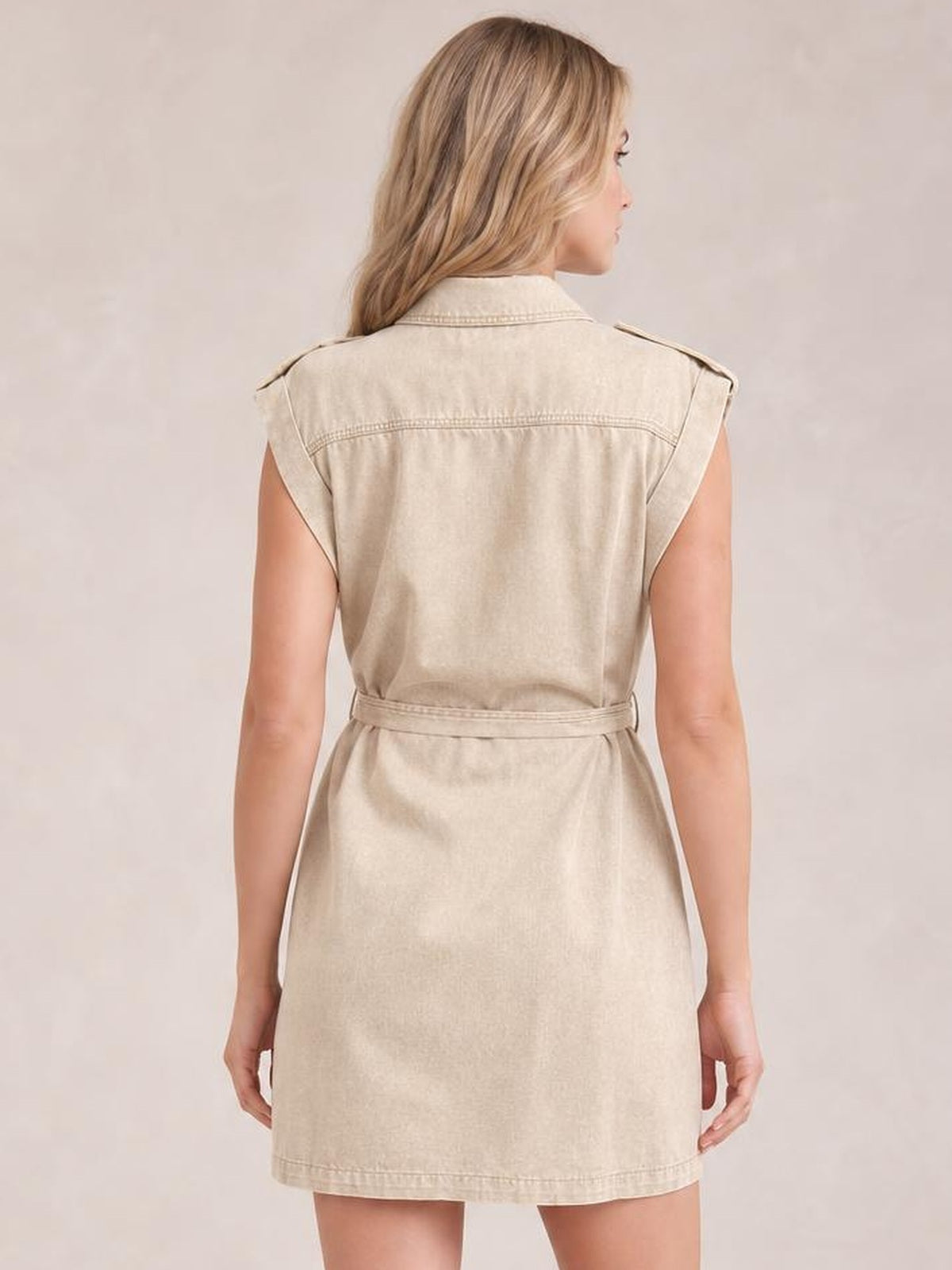 Model shown from behind wearing a light beige utility-style denim mini dress with a collared neckline, sleeveless cut, waist tie belt, seamed construction, and straight silhouette.