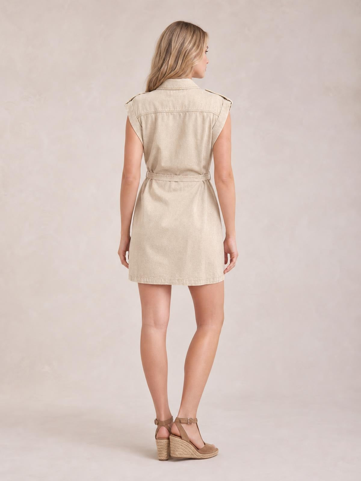 Model shown from the back in a light beige denim mini dress with a collared neckline, sleeveless utility styling, button front, and tied waist belt.