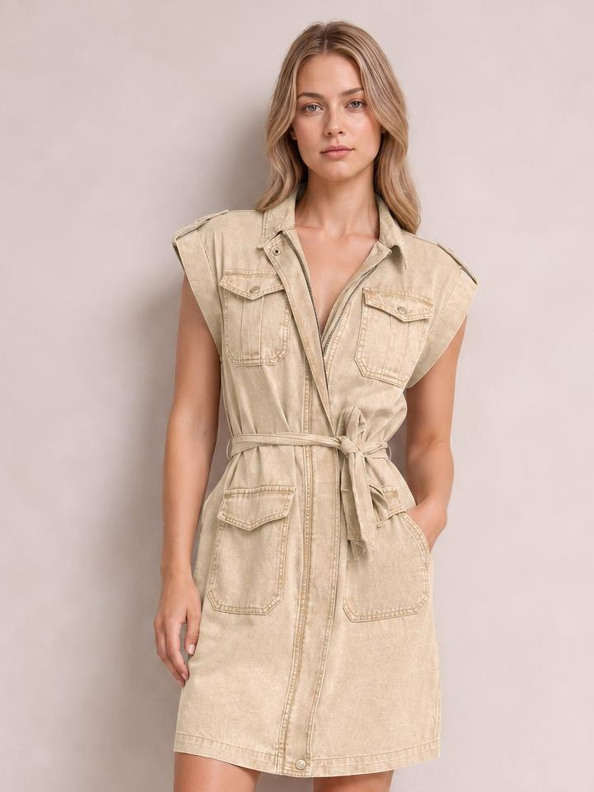 Model poses in a light beige denim mini dress with a collared neckline, sleeveless utility style, button front, multiple flap pockets, and tied waist belt.
