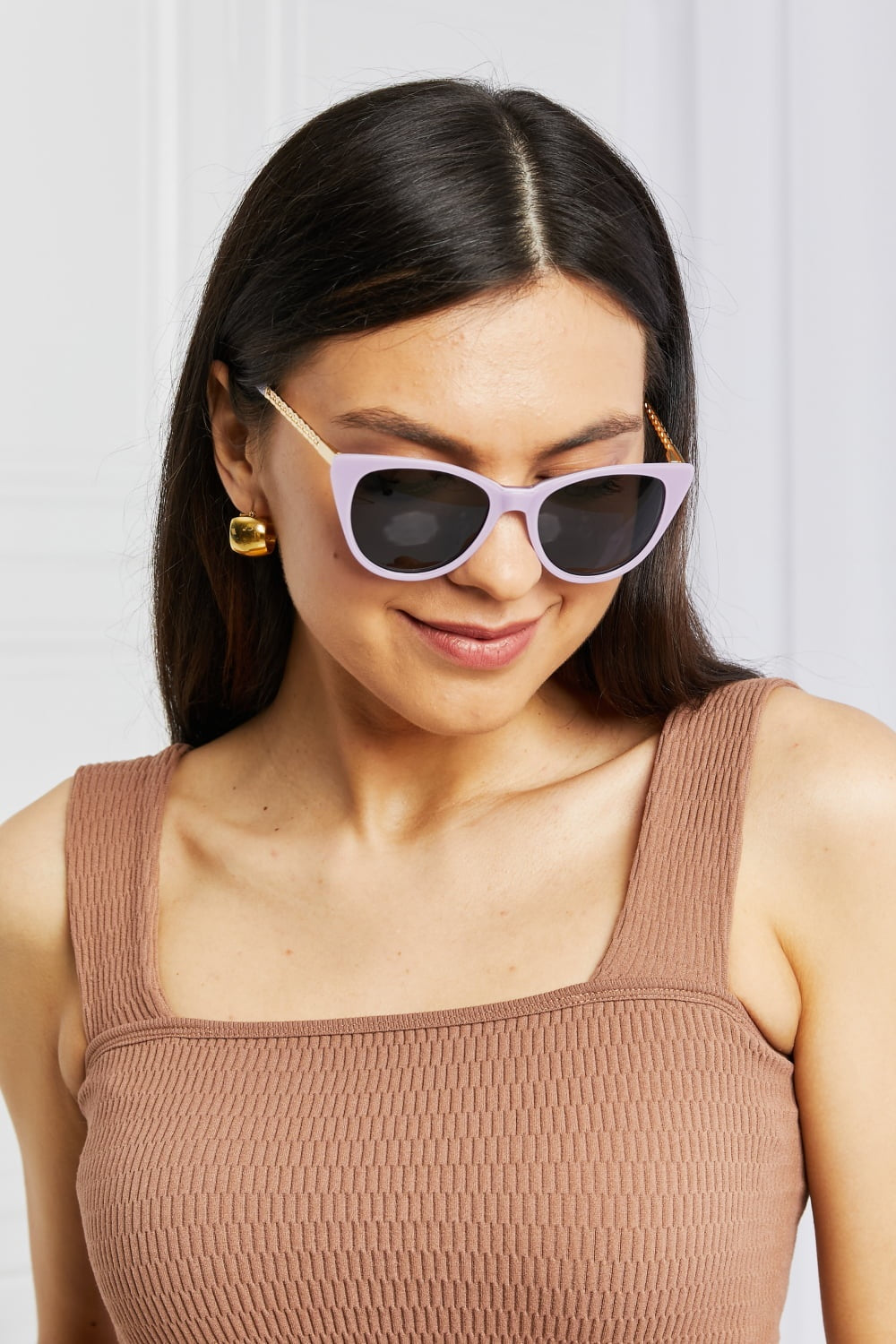 A smiling model looks downward while wearing lilac acetate cat-eye sunglasses with dark lenses, softly upswept frames, and gold-tone metal temple detailing.