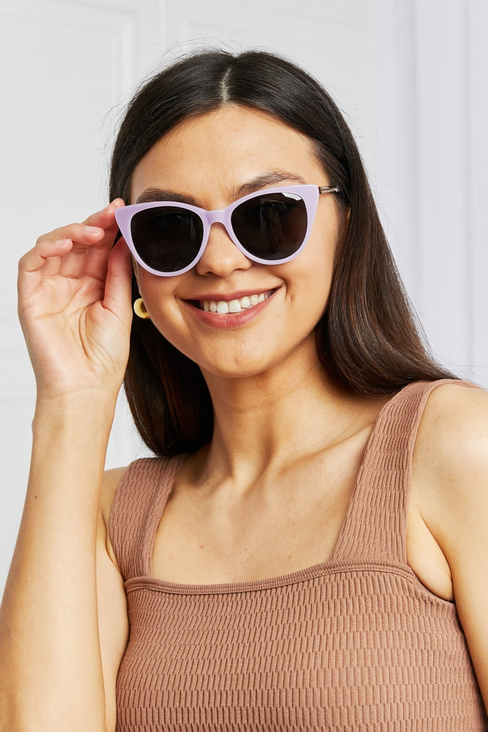Smiling model adjusts lavender acetate cat-eye sunglasses with dark lenses and metal-acetate temples, showcasing their softly upswept, face-framing silhouette.