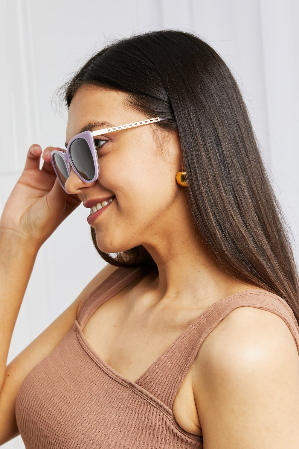A smiling model adjusts lavender cat-eye acetate sunglasses with dark lenses and decorative metal-acetate temples, showing their softly upswept silhouette.