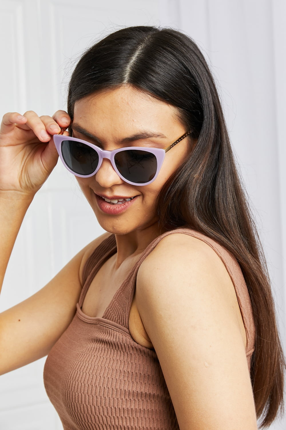 A smiling model adjusts lavender acetate cat-eye sunglasses with dark lenses and metal-acetate temples, wearing a brown ribbed tank top in a close-up pose.