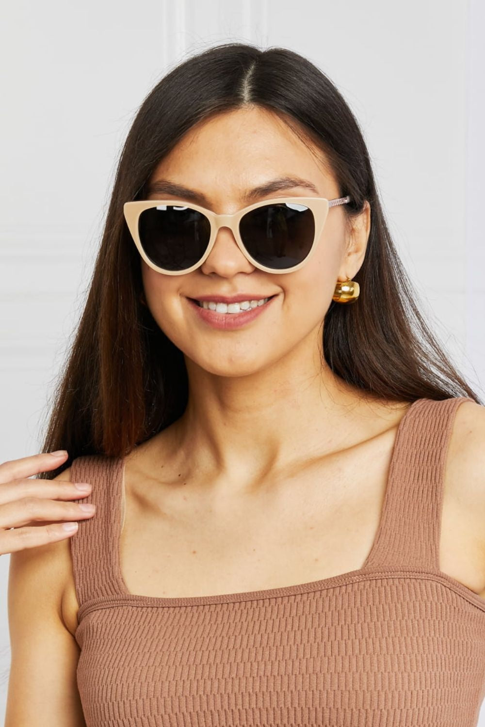 Smiling model touches her hair while wearing cream cat-eye acetate sunglasses with dark lenses and a taupe ribbed square-neck sleeveless top.