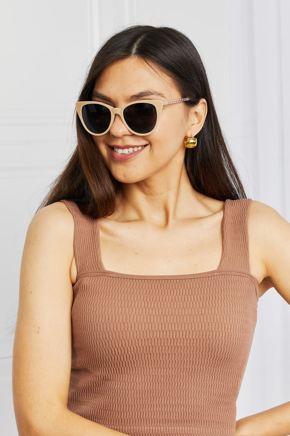 Smiling model wears beige acetate cat-eye sunglasses with dark lenses and mixed metal-acetate temples, styled with a brown square-neck sleeveless top.