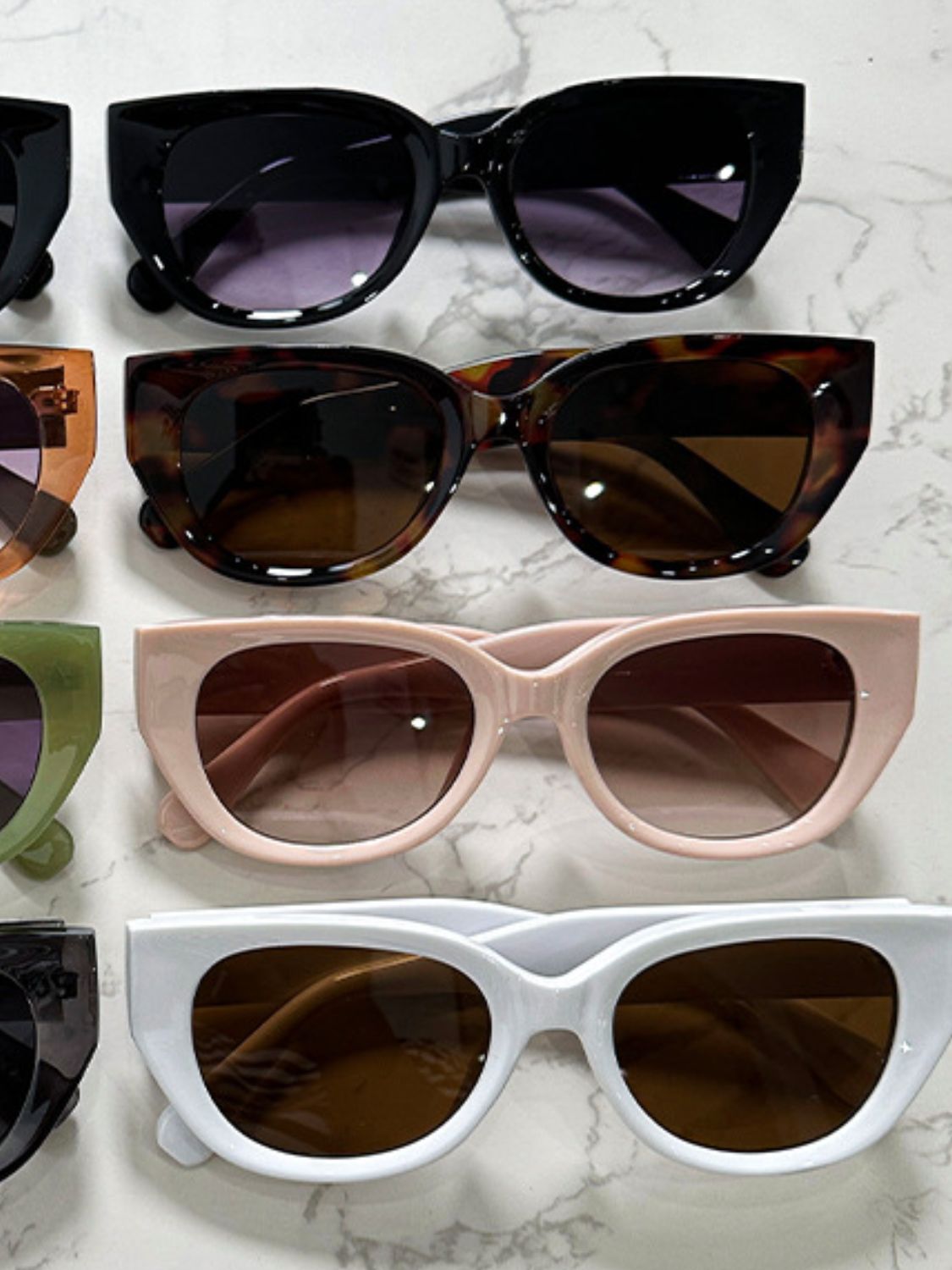 Rows of cat-eye polycarbonate sunglasses in black, tortoiseshell, blush, and white feature rounded lenses, bold frames, and softly upswept corners.