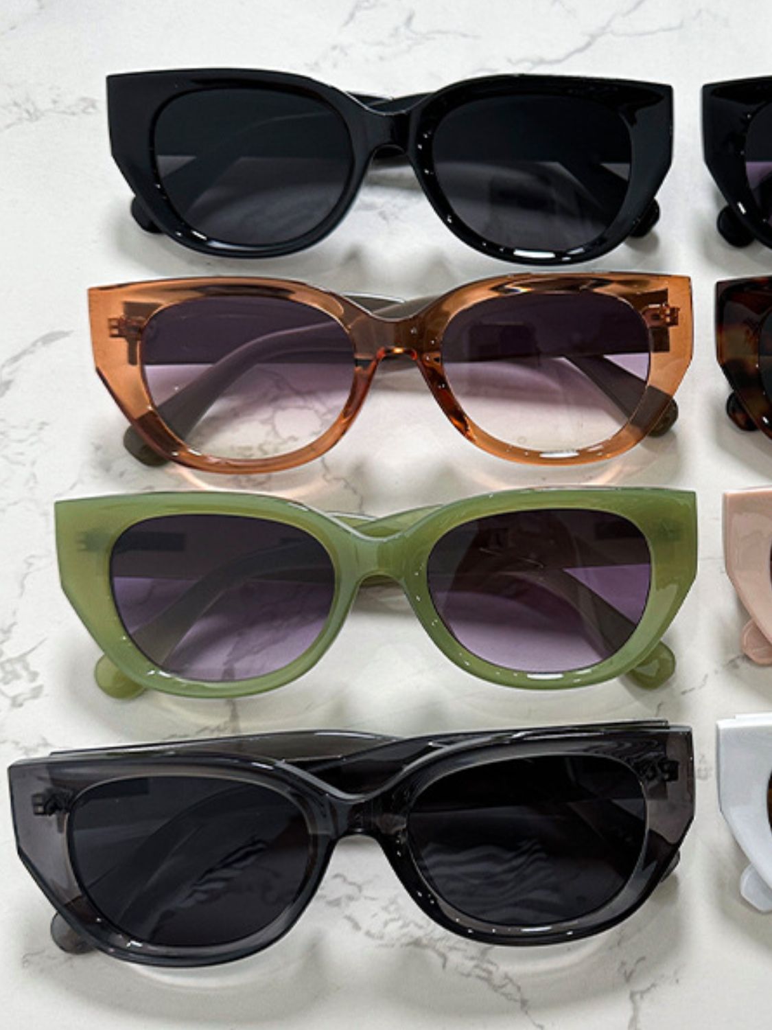 Assorted polycarbonate cat-eye sunglasses feature bold glossy frames in black, tortoiseshell brown, olive green, and dark charcoal with rounded lenses and upswept corners.