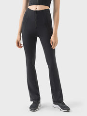 A model stands in black high-waist active pants with a front zipper, contoured seam detailing, and a sleek, fitted straight-leg silhouette.