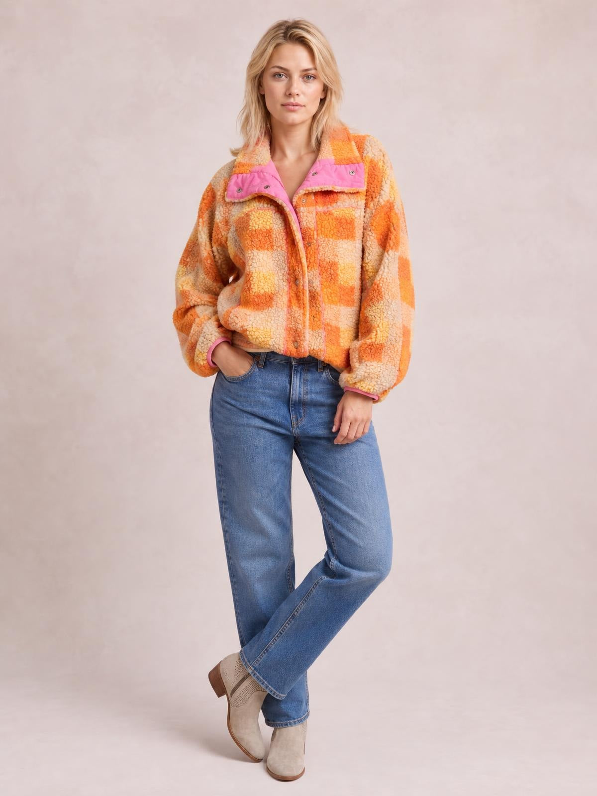 A model poses in an orange and cream checkered sherpa jacket with long sleeves, pink-trimmed chest pockets, button closure, and a relaxed silhouette.