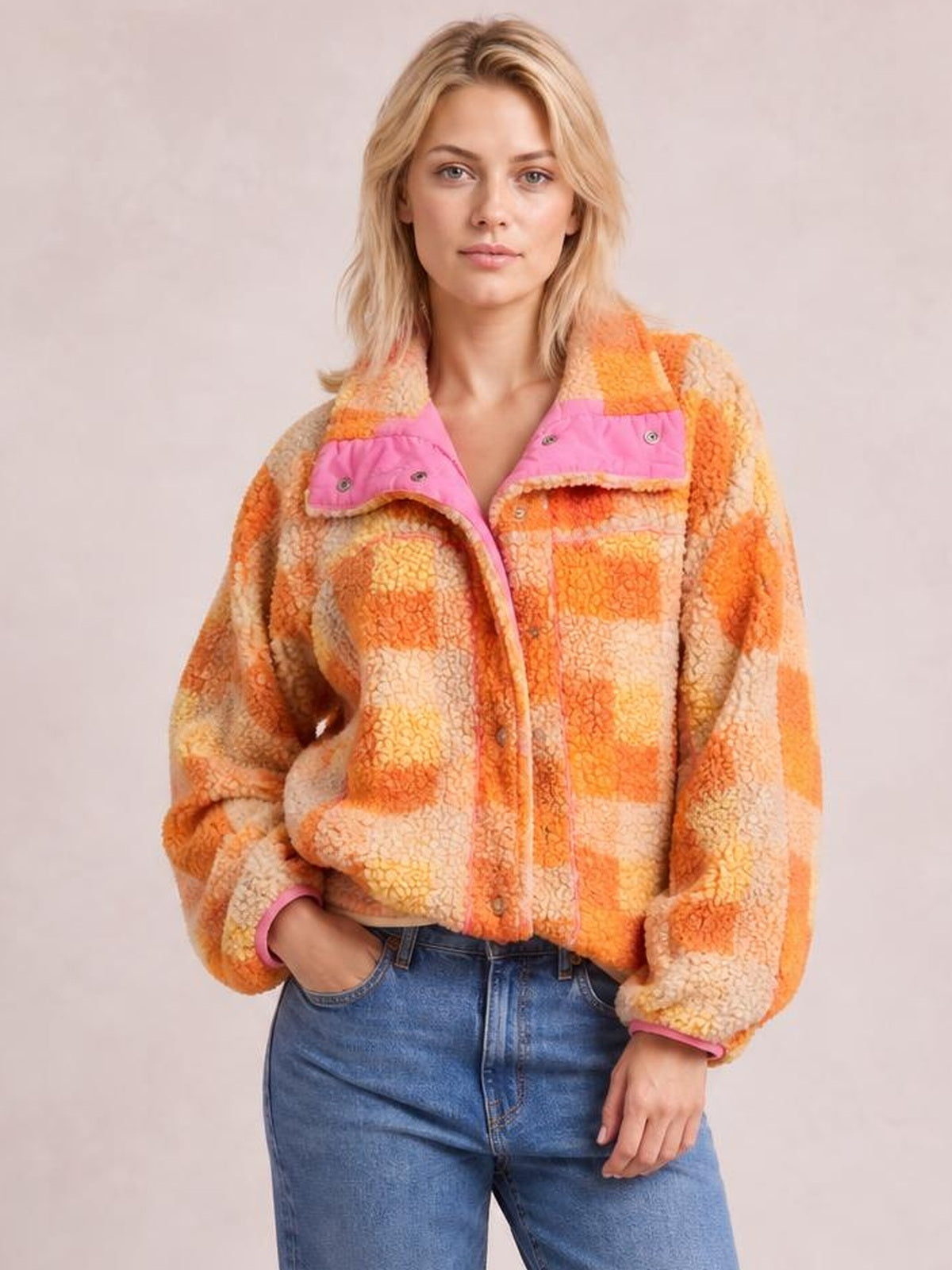 A model poses with one hand in her pocket, wearing an orange-and-cream checkered sherpa jacket with long sleeves, snap front, and patch pockets.