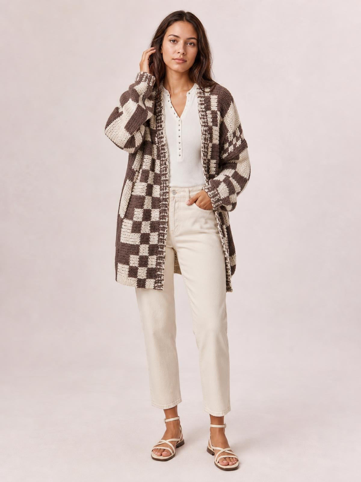 A model touches her hair with one hand and rests the other in a pocket, wearing a brown-and-cream checkered open-front cardigan with long sleeves and a relaxed silhouette.