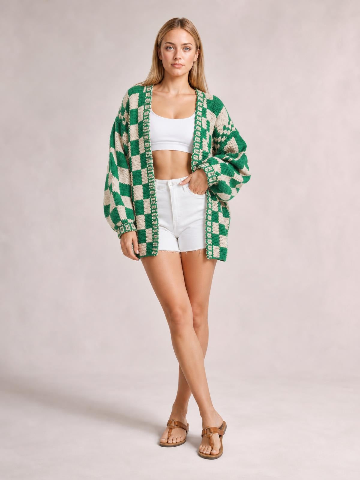 A model poses with crossed legs in a green-and-cream checkered open-front long-sleeve cardigan with a relaxed oversized silhouette and ribbed edges.