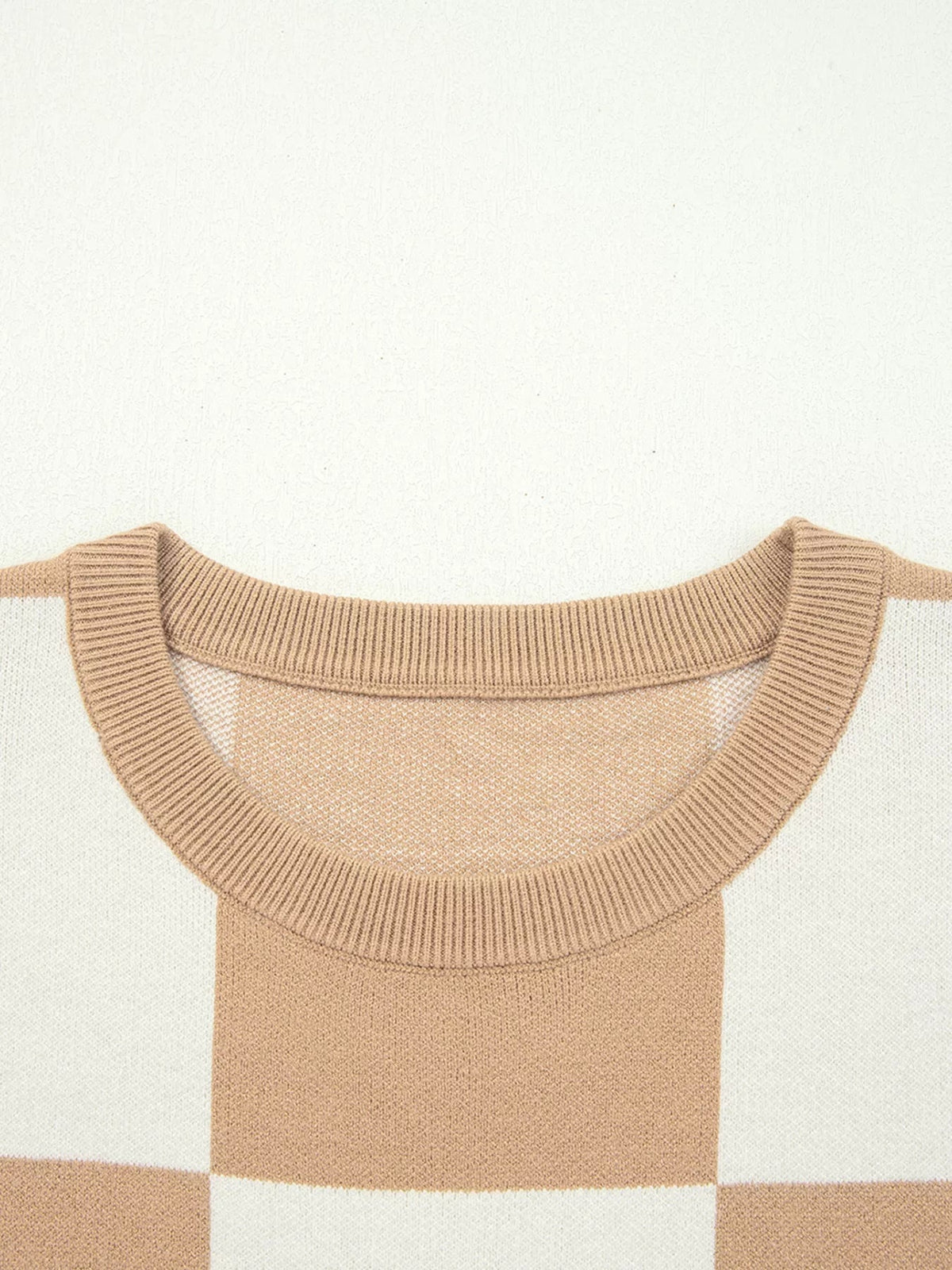 Close-up of a tan checkered sweater showing its loose knit construction, ribbed crew neckline, drop shoulder seams, and half-sleeve design.