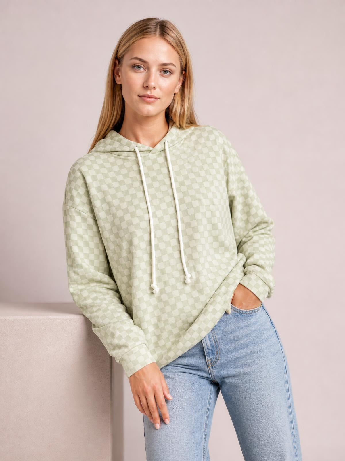 A model wears a pale green checkered hoodie with a relaxed silhouette, long sleeves, drawstring hood, and kangaroo pocket while posing with one hand tucked inside.