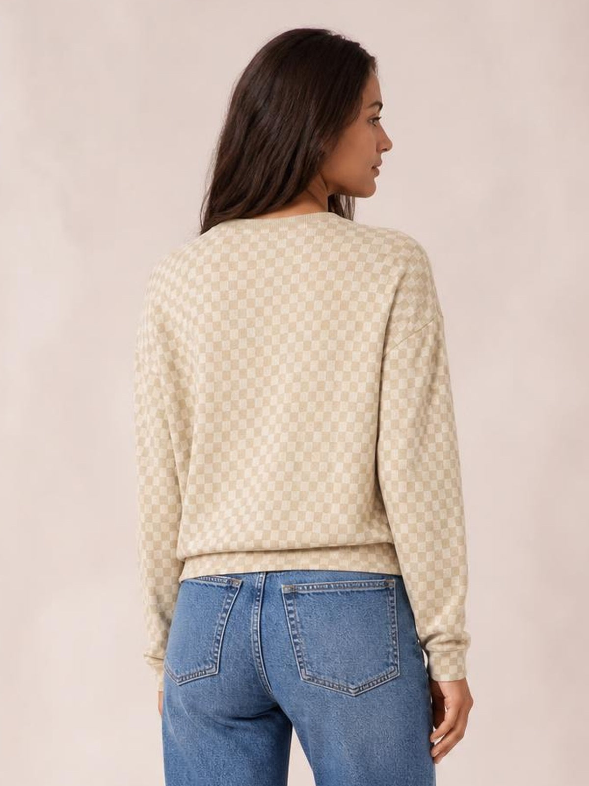 A model poses from behind in a cream checkered hoodie with long sleeves, a relaxed straight silhouette, and a gently gathered hem above blue jeans.