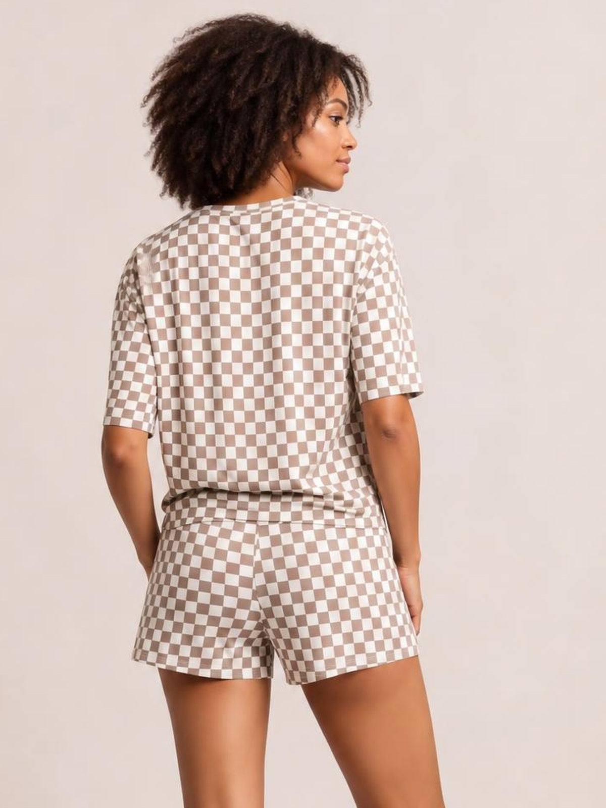 A model poses from behind in a beige-and-white checkered round-neck top with short sleeves and matching relaxed-fit drawstring shorts.