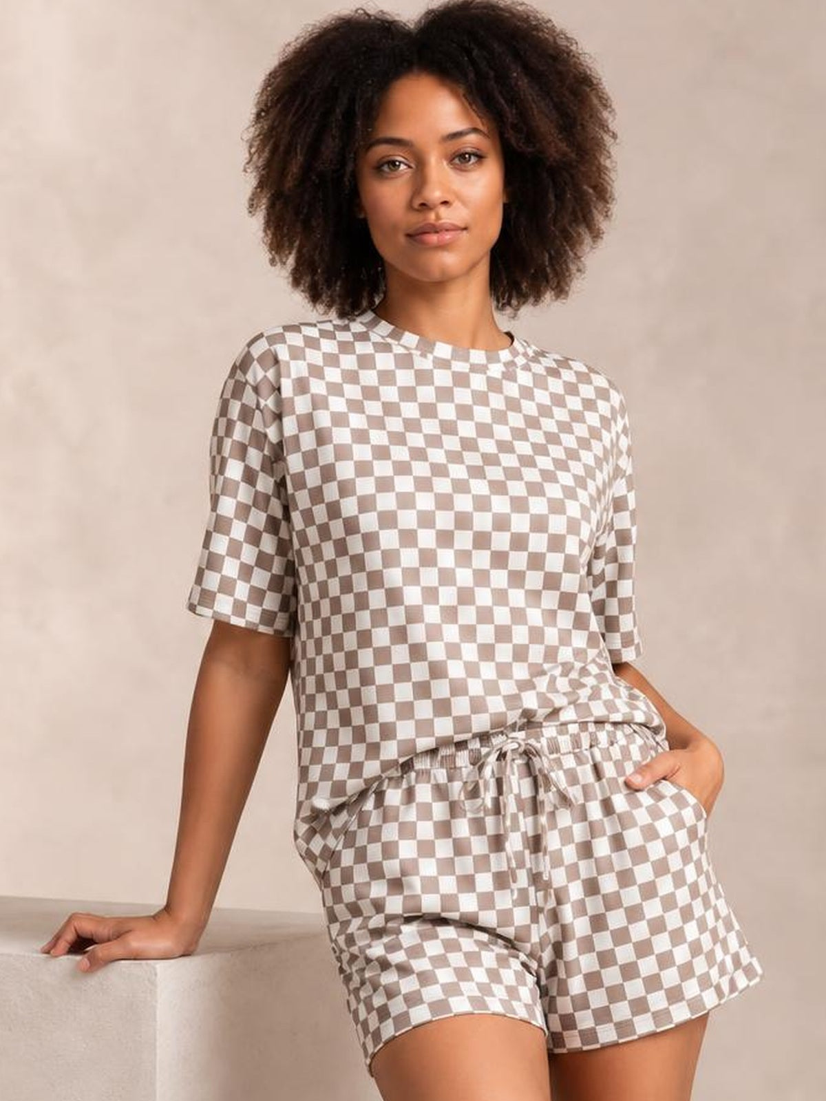 Model leans against a surface wearing a beige-and-white checkered round neck top with short sleeves and matching drawstring shorts in a relaxed silhouette.