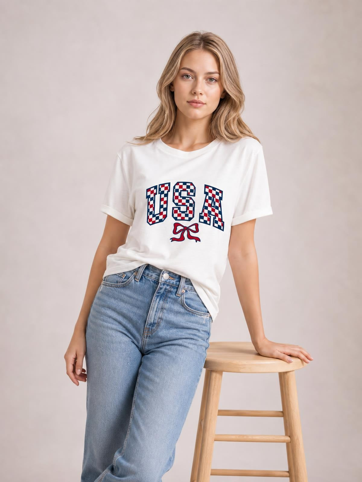 Model leans against a stool wearing a white round neck tee with rolled sleeves, a checkered USA graphic, and a relaxed straight silhouette.