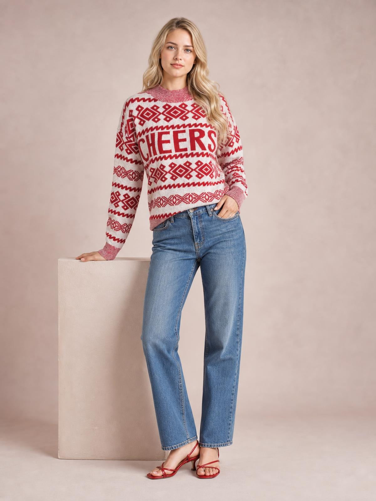 A model leans against a pedestal wearing a red-and-white Christmas sweater with a round neck, long sleeves, geometric knit pattern, and bold CHEERS lettering.
