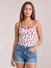 Model wearing a white cherry print tank with a square neckline, red trim, wide shoulder straps, and a fitted silhouette, posed with one hand in denim shorts pocket.