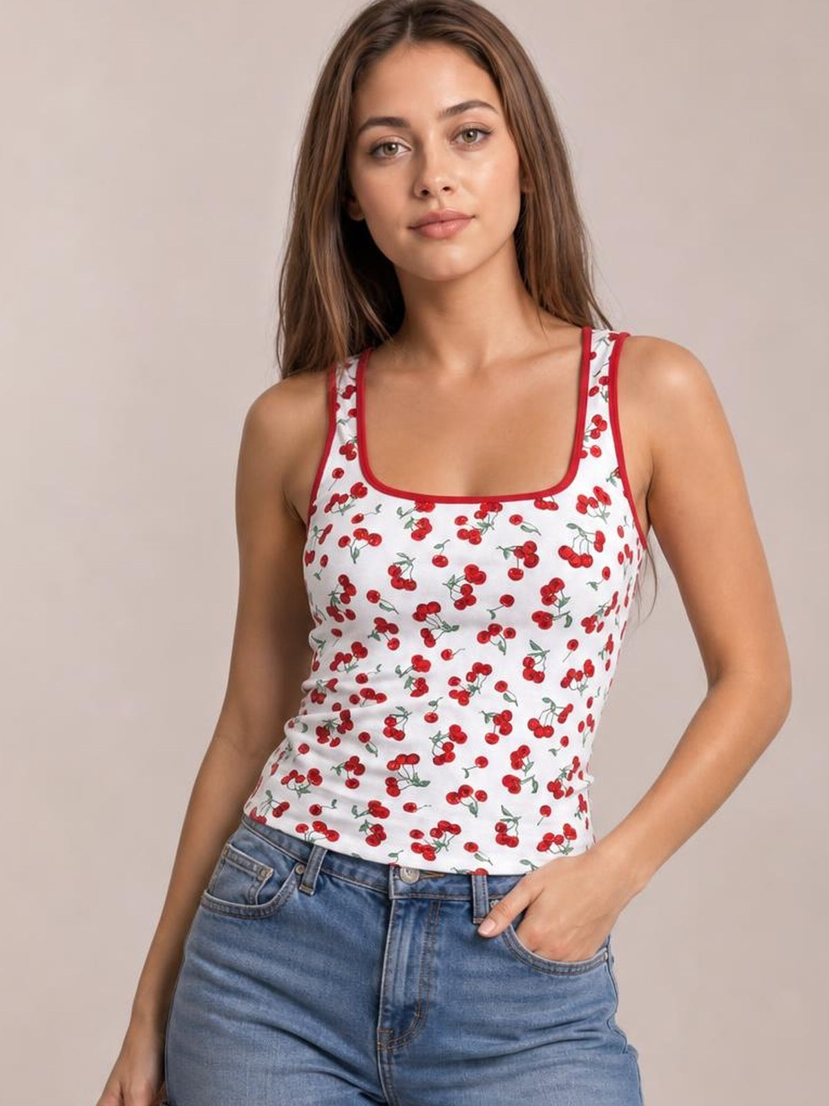 Model wears a fitted white cherry print tank with a square neckline, red trim, and wide shoulder straps while standing with one hand in her jeans pocket.