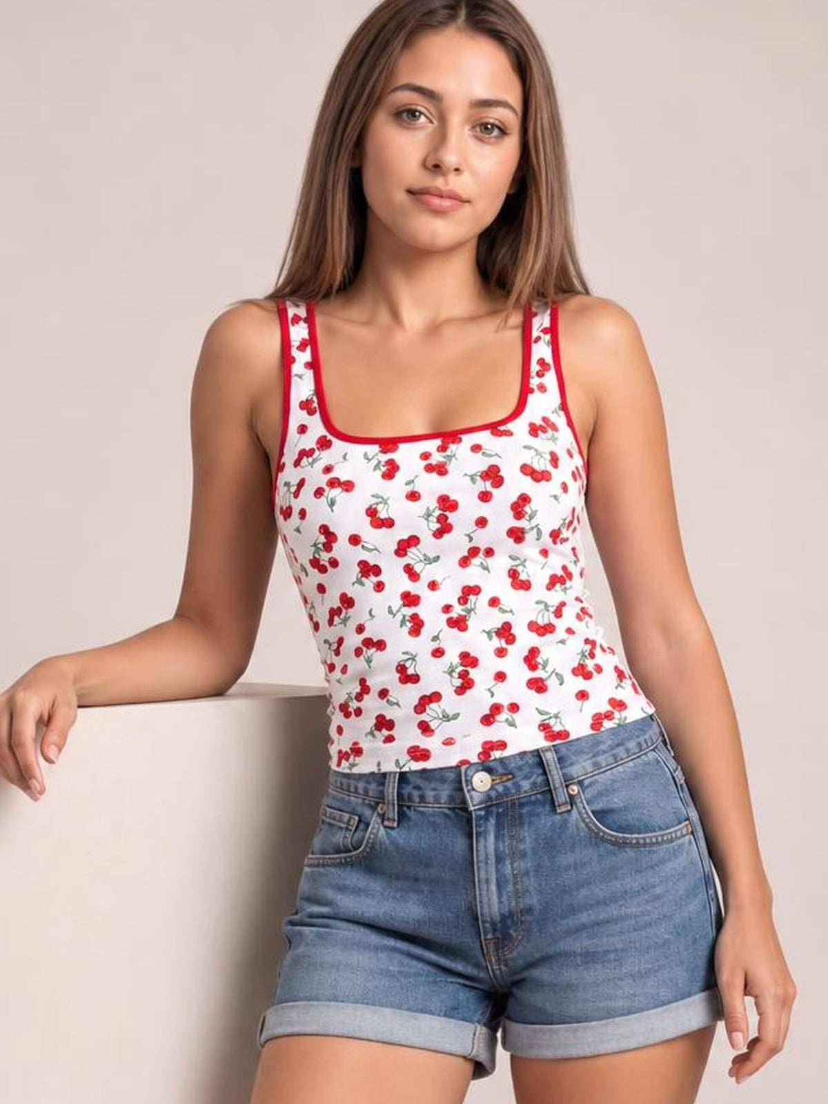 Model wears a fitted white cherry print square neck tank with red trim and wide straps, paired with denim shorts while resting one arm beside her.