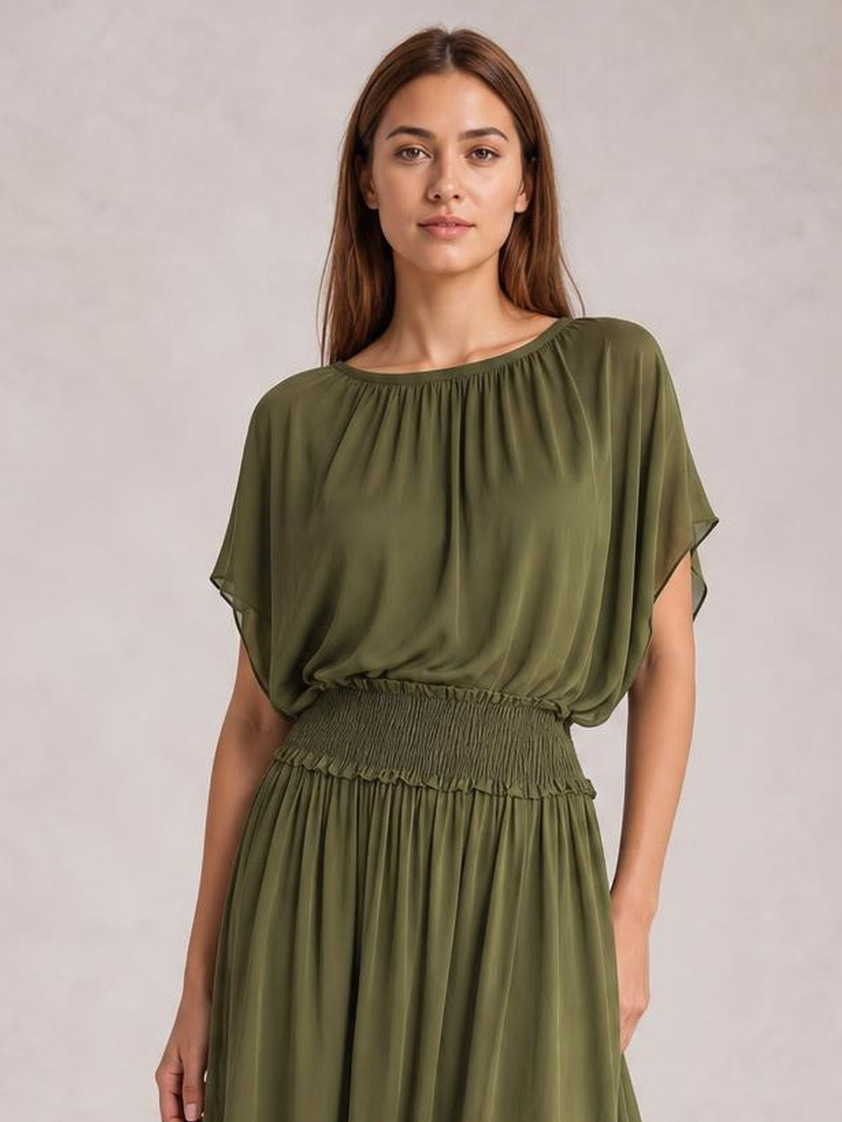 A model stands facing forward in an olive green chiffon maxi dress with a gathered neckline, flutter sleeves, smocked waist, and flowing pleated skirt.