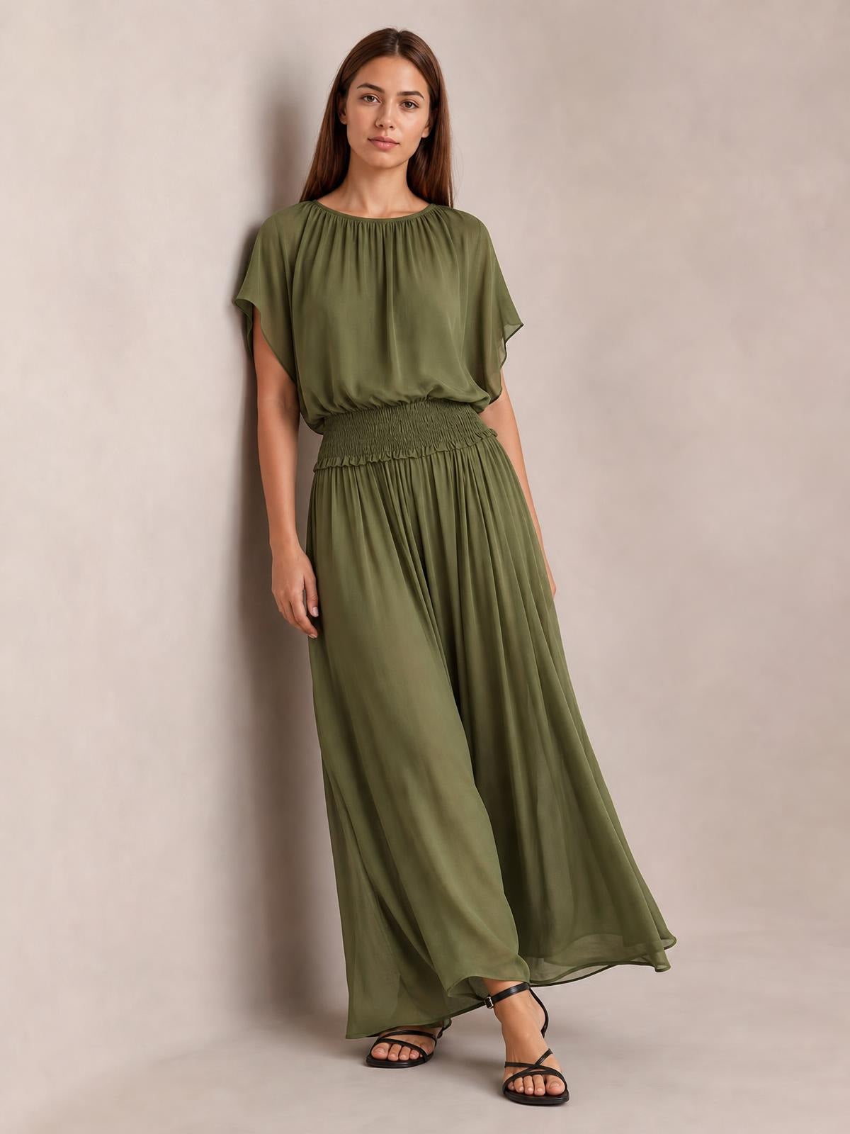 Model wears an olive green chiffon maxi dress with a smocked waist, gathered round neckline, flutter sleeves, and a flowing full-length silhouette.