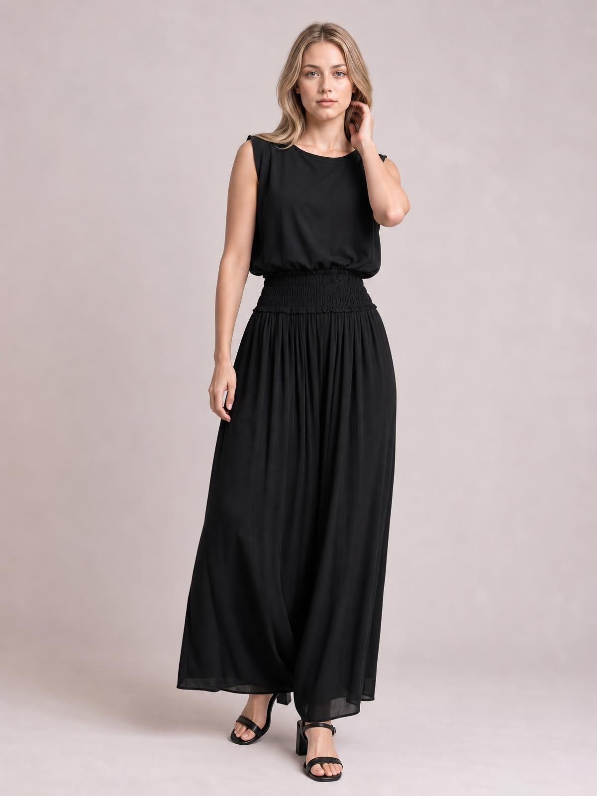 A model poses in a black chiffon maxi dress with a sleeveless round-neck bodice, smocked waist, and flowing pleated skirt.