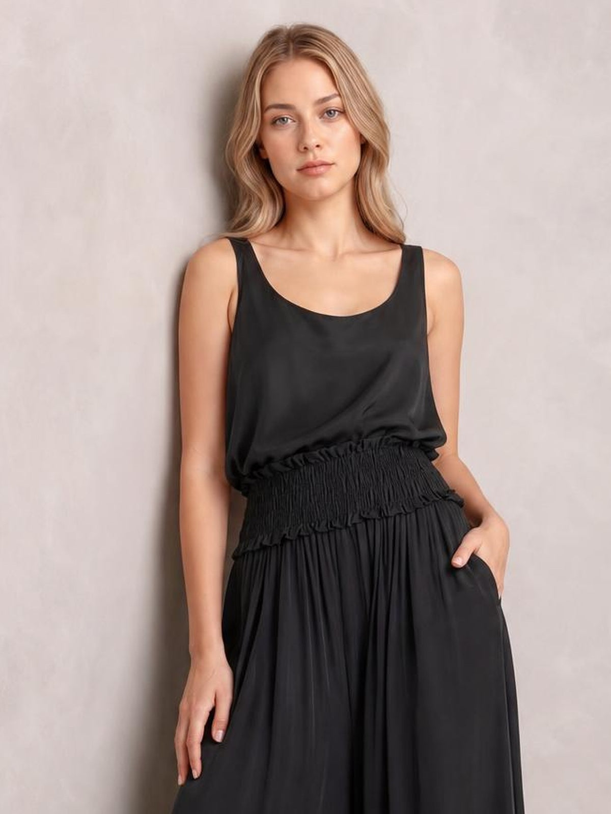 A model wears a black chiffon maxi dress with a sleeveless scoop neckline, smocked waist, flowing gathered skirt, and one hand in a side pocket.