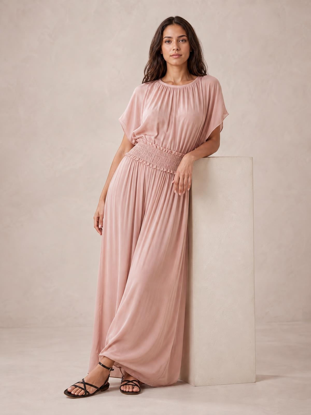 Model leans against a block wearing a blush pink chiffon maxi dress with a gathered neckline, flutter sleeves, smocked waist, and flowing floor-length silhouette.