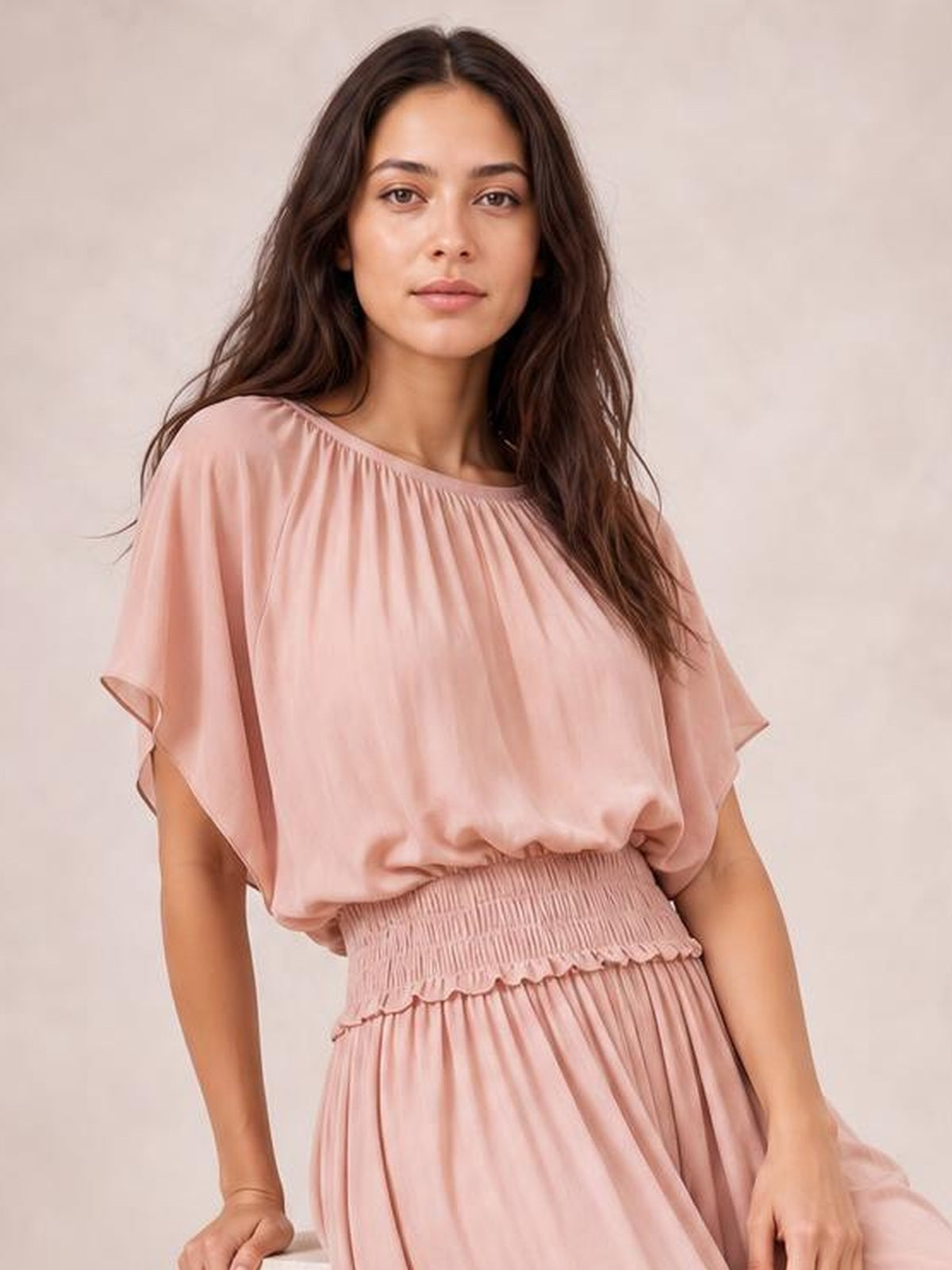 A model poses in a blush pink chiffon maxi dress with a wide neckline, flutter sleeves, smocked waist, and flowing pleated skirt.