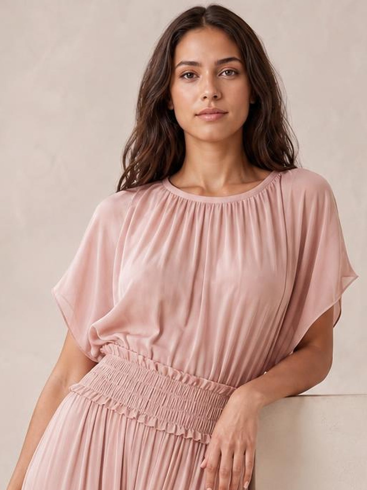 Model leans against a ledge wearing a blush pink chiffon maxi dress with a gathered neckline, flutter sleeves, and smocked waist.