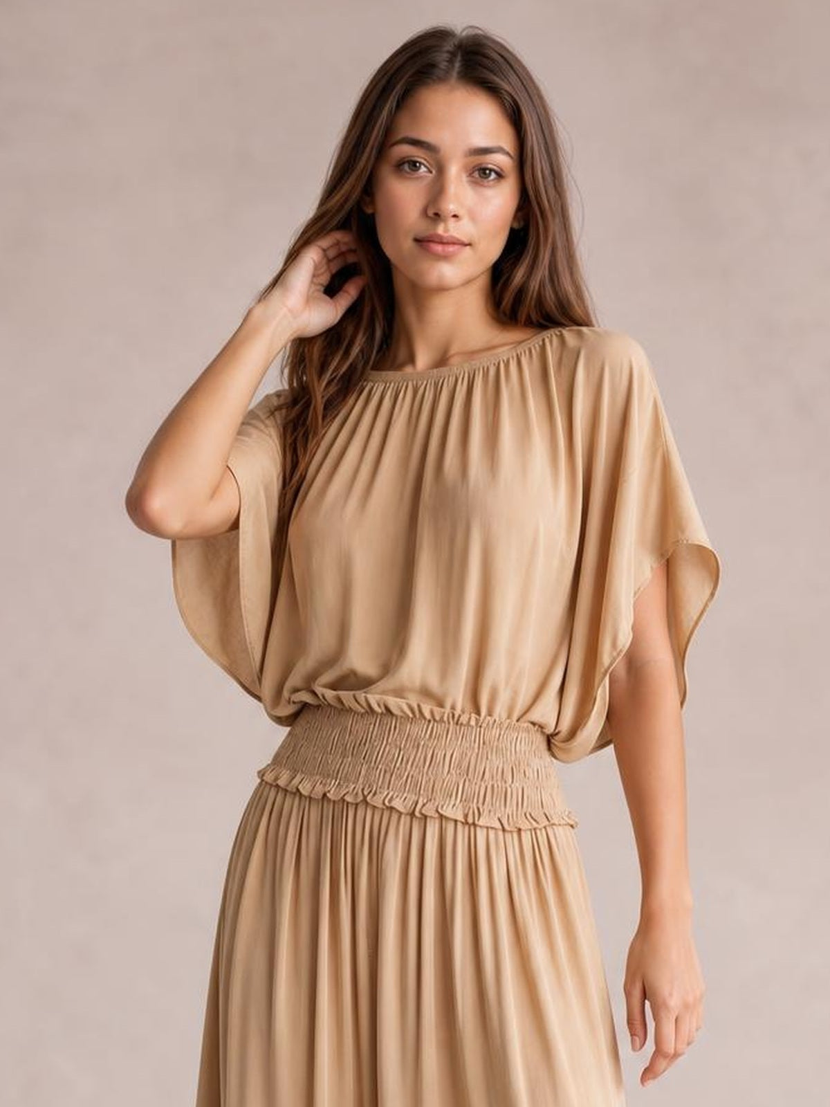 Model poses with one hand in her hair wearing a beige chiffon maxi dress with a gathered neckline, flutter sleeves, smocked waist, and flowing pleated skirt.