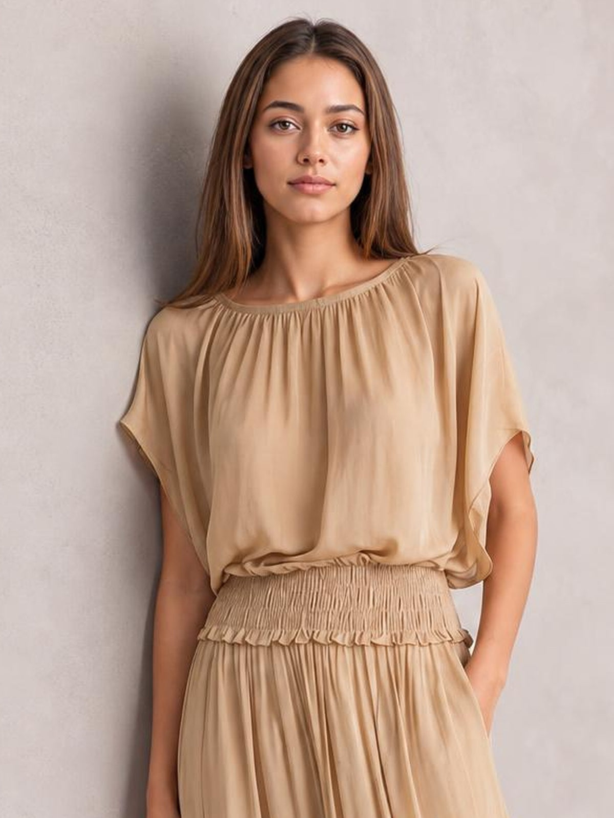 A model poses in a beige chiffon maxi dress with a gathered round neckline, flutter sleeves, smocked waist, and flowing pleated skirt.